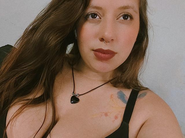 ArenaPeeach with brown hair, nose ring, and black lipstick, wearing a black tank top, showing her tits and tattoo, necklace with heart pendant, looking at camera, soft smile.