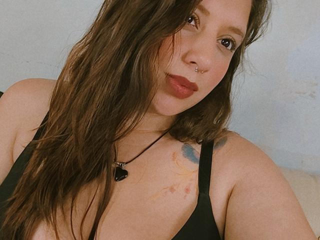 ArenaPeeach met lang bruin haar, een neusring en rode lippenstift, draagt een zwarte tanktop, toont een tatoeage op haar rechter schouder en een zwarte hartketting..