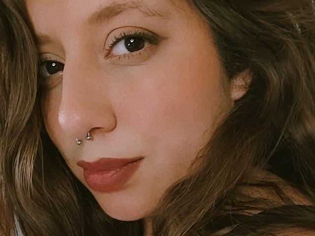 ArenaPeeach hat ein Nasenpiercing mit einem kleinen silbernen Ring in ihrem rechten Nasenloch, und ihr braunes, welliges Haar ist teilweise sichtbar. Sie trägt einen dezenten roten Lippenstift..