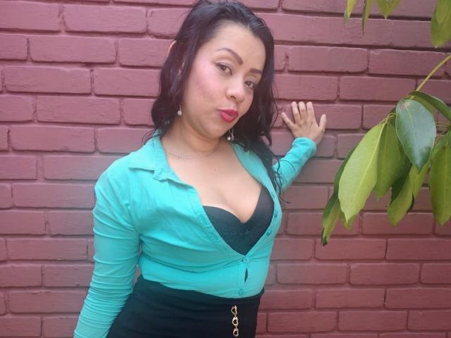 ThaliaSaenz with puffy red lips, green cardigan, black bra showing her big tits, black skirt, and long dark hair, leaning against a red brick wall, hand touching wall.