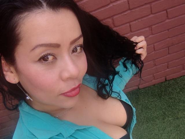 Close-up selfie of ThaliaSaenz with fair skin, dark wavy hair, and brown eyes. She's wearing a turquoise open shirt, revealing a black bra that shows her cleavage. ThaliaSaenz has red lipstick, subtle eye makeup, and small silver earrings. She's holding her hair with her right hand. The background features a red brick wall and green grass. The image focuses on her upper body and face, with a slight upward angle. She has a confident expression.