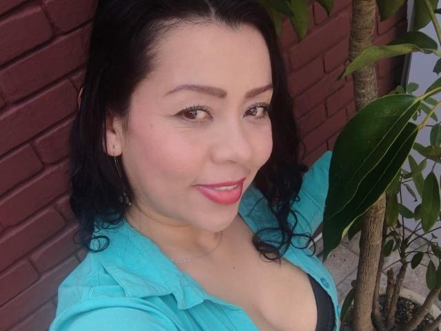 ThaliaSaenz has dark hair, pink lipstick, and is smiling with her tits slightly visible in a black top under a blue shirt. She's standing next to a green plant.