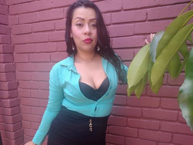 ThaliaSaenz stands against a red brick wall, leaning slightly to the right. She has long, dark, wavy hair and wears a teal, partially unbuttoned cardigan over a black bra that highlights her medium-sized breasts. She has fair skin and bright red lipstick. She wears a black skirt with a gold zipper, and her right hand touches a green leaf. The image is clear and focused.