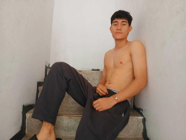 JayOrange, shirtless, with small tits, sits on gray stairs, wearing dark pants, barefoot, black hair, bracelet on right wrist, neutral expression, white walls, corner of stairs.