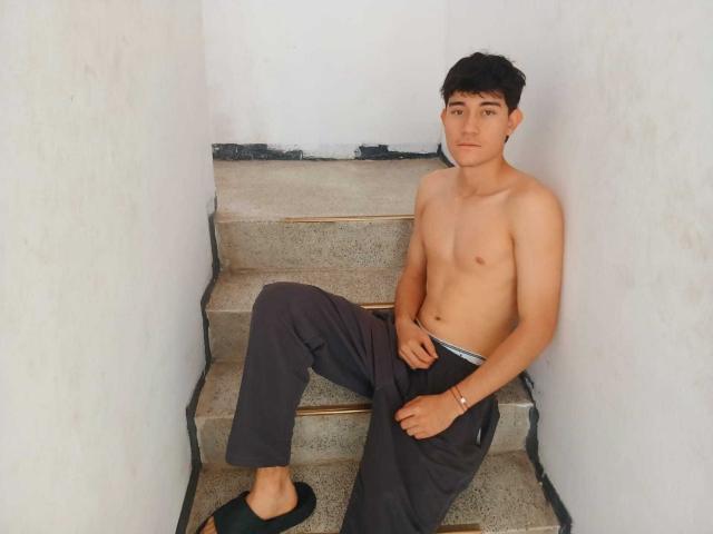 JayOrange, shirtless with dark pants, black slippers, and a white underwear waistband, sits on concrete stairs, right hand on pants, left hand on knee, looking at camera.