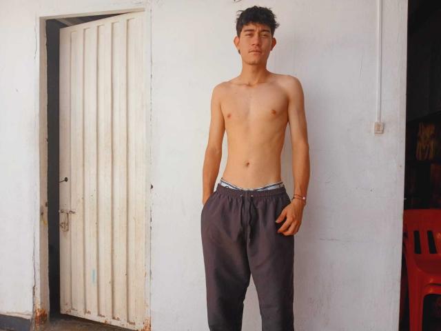 Shirtless JayOrange with a slim, toned build, dark curly hair, and hands in loose grey pants, standing against a white wall, looking confident.
