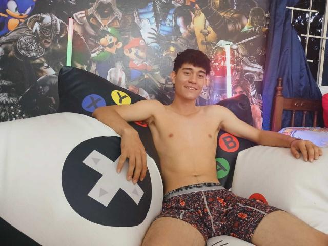 JayOrange, shirtless, with brown hair, smiling, wearing colorful boxer shorts, sitting on a white couch with video game pillows, in a room with a video game-themed wall.