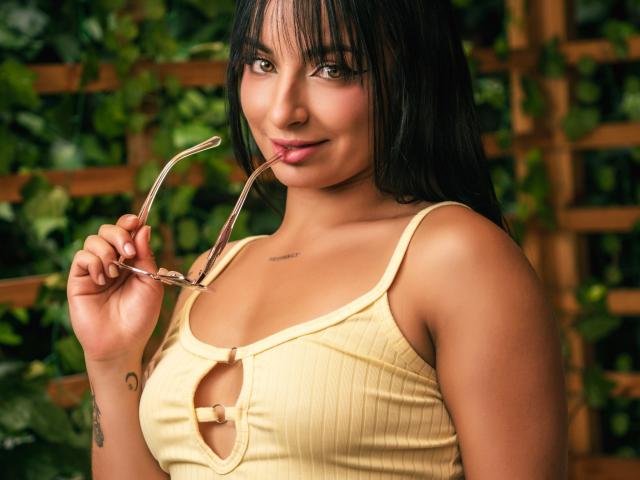 IsadoraGallego with dark hair, holding glasses in her mouth, wearing a yellow ribbed top with a cutout, showing her tits, smiling, tattoo on her right arm.