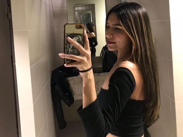 AfroditaWild with long brown hair, red lipstick, black off-shoulder top, holding phone in mirror selfie, showing her back and small tits, in bathroom with beige tiles.