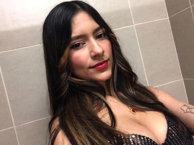 AfroditaWild has long dark brown hair, red lipstick, and is wearing a sparkly black top showing her large tits. She's looking at the camera with a slight smile.