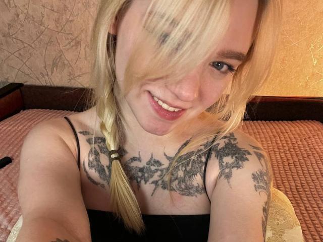 EnchantingEyes with blonde hair in a braid, smiling, showing tattoos on her chest and arms, wearing a black strap top, in a selfie. Her tits are visible.