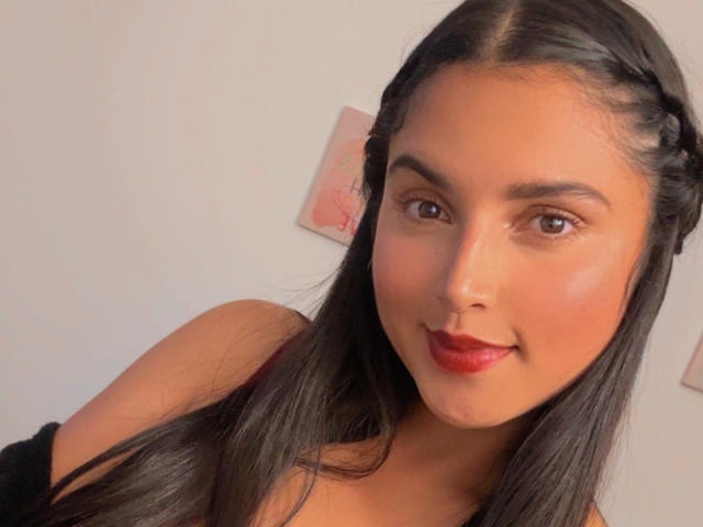 VeronicaFire69 with long black hair in braids, brown eyes, red lipstick, and a slight smile, looking at the camera. She's wearing a black top, showing her shoulders.