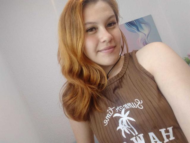 TaylorCarpert with light brown hair, smiling, wearing a brown sleeveless top, showing her tits, fair skin, and a small black necklace, standing in a bright room.