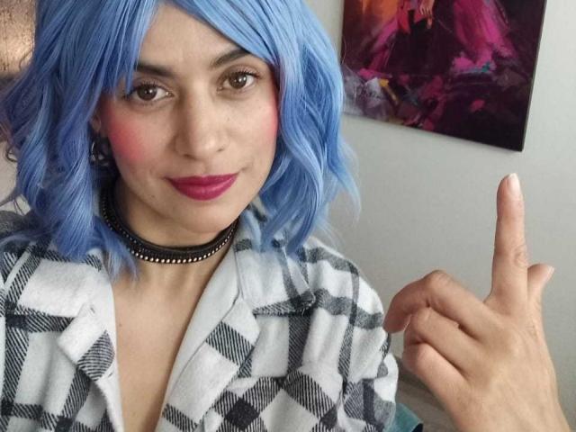 LilaaSalmon with blue hair, pink blush, purple lipstick, black choker, checkered shirt, and finger pointing, looking at camera. Boobs not visible. 

 