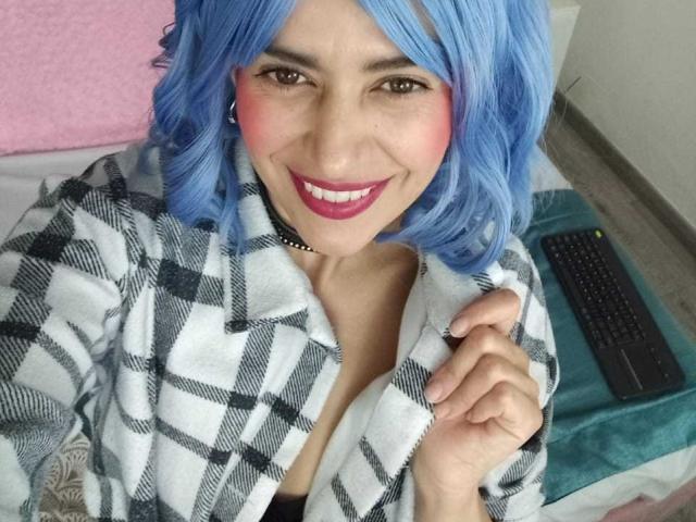 LilaaSalmon with blue hair, pink blush, and pink lipstick, smiling at the camera, wearing a checkered shirt, showing her tits slightly, holding her shirt.