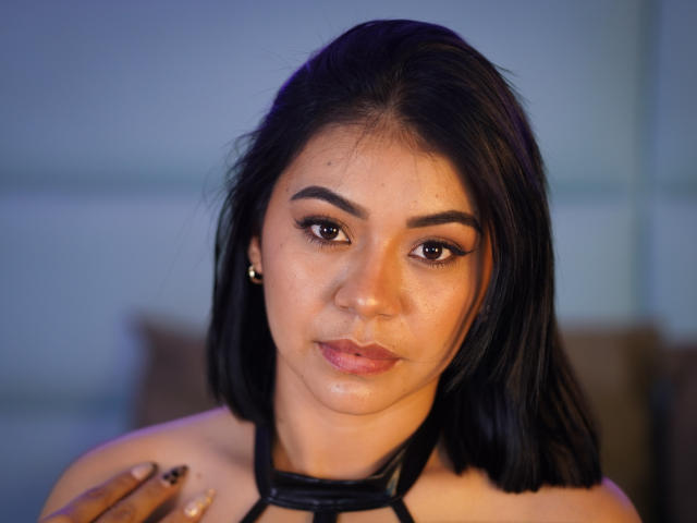 AngelVega has medium brown skin, black shoulder-length hair, and is wearing a black halter top, showing off her ass slightly. Her makeup is subtle with winged eyeliner.