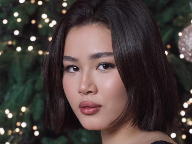 A close-up portrait of ArianaFlower. She has a medium skin tone, straight black shoulder-length hair, and full pink lips. Her dark eyebrows are slightly arched, and she has subtle eyeliner. Her expression is neutral, slightly pouty. The background is blurred with small, warm, white lights. The composition centers on her face, highlighting her smooth, flawless skin. The lighting is even, emphasizing her natural beauty.