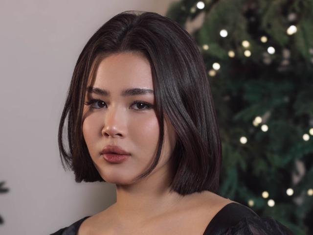 ArianaFlower has a medium skin tone and a sharp, bob-style haircut with straight, dark brown hair. She has thick, dark eyebrows and is wearing subtle makeup with dark eyeliner, long eyelashes, and a muted pink lipstick. She looks directly at the camera with a neutral expression. She is wearing a black, lacey top. A blurred, decorated Christmas tree is in the background. The image is focused on her face and upper torso.