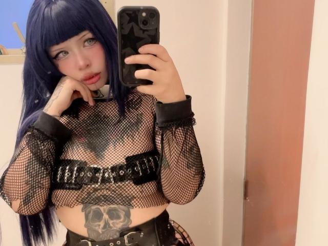 StarWitch with long blue hair, wearing a fishnet top, black cuffs, and a skull tattoo on her belly, taking a mirror selfie with a phone, showing off her tits.