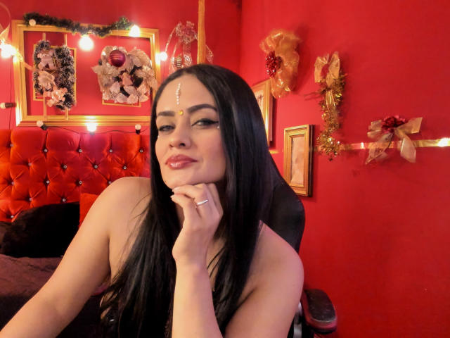 AnissaAli with long black hair, red lipstick, and a facial dot, resting her chin on her hand, wearing a black strapless top, in a red room with Christmas decor.