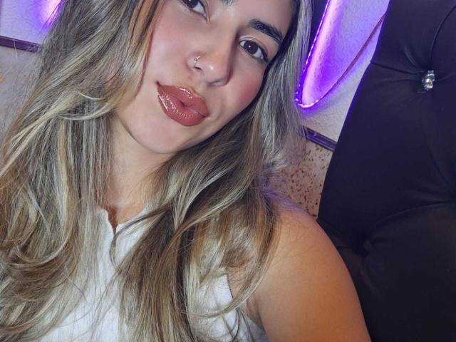 ScarlettKhan has long, wavy, blonde hair, shiny pink lips, a nose ring, and is wearing a white top, with purple neon lights in the background.