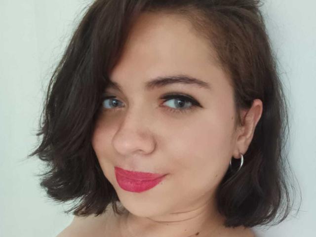 LilaSquirt has short brown hair, blue eyes, and red lipstick. She's smiling slightly with a hoop earring in her left ear. Her boobs are not visible.