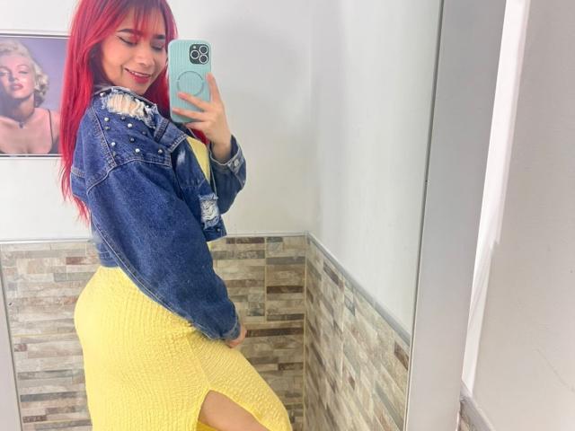 MelannyTaylorr with red hair, smiling, wearing a ripped blue denim jacket and yellow skirt, showing her ass in a mirror selfie. Her phone has a teal case.