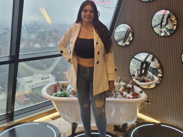 katherinLatin stands in a cafe, wearing a beige coat, black strapless top, and ripped blue jeans, with a smiling face, long brown hair, and a cute ass.