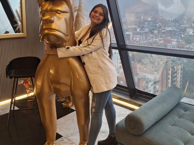 KatherinLatin with long brown hair, smiling, standing next to a shiny gold horse statue, wearing a white coat and blue jeans, in a modern room with a city view.