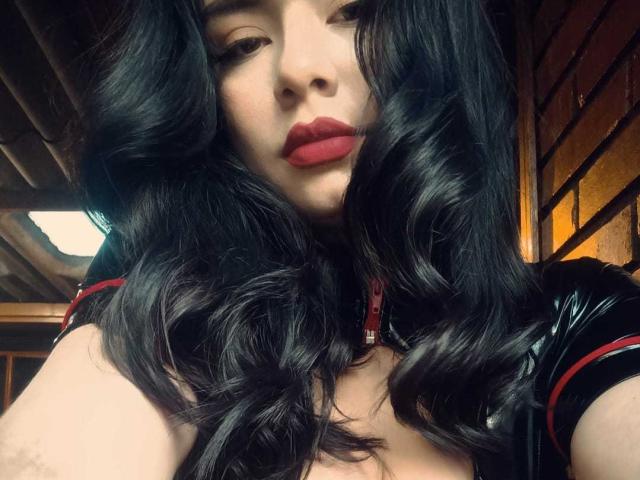 RoseAnna with wavy black hair, red lipstick, and glossy black top, showing her tits, looking down at the camera, in a wooden room.