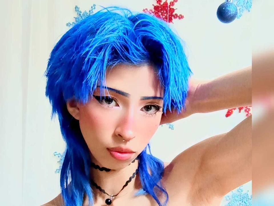TinaRoberts69 has bright blue hair, a nose ring, black eyeliner, and a black choker. Her arm is raised, showing a bare, toned, and muscular upper body.