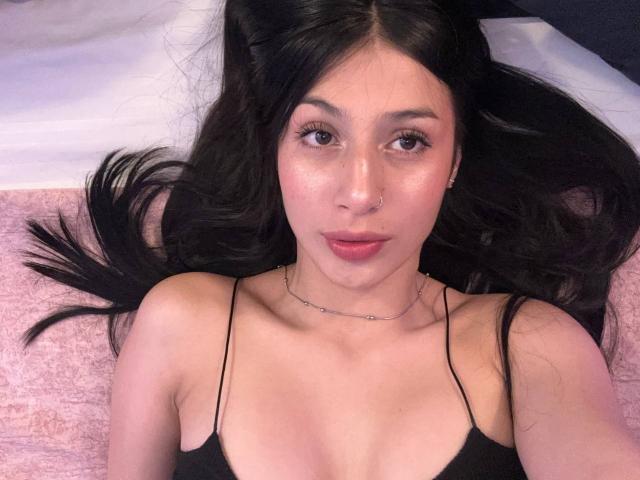 SophieBeaumont with long black hair, looking up, wearing a black spaghetti strap top, showing her small tits, nose ring, and subtle makeup, lying on a pink surface.