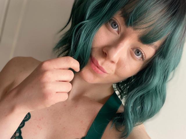 JessieFire-hot with teal hair, fair skin, and blue eyes, holding a green strap against her chest, slightly biting her lip, looking directly at the camera.