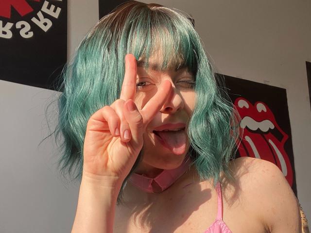 JessieFire-hot with teal hair, pink choker, and pink top, sticking out tongue, making a "peace" sign with right hand, sunlight on face, looking cute and playful.