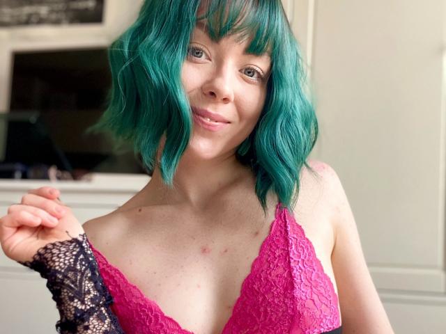 JessieFire-hot with teal hair, pink lace bra, and black lace glove, smiling, showing small tits, light skin, slight acne, and right hand touching her hair.