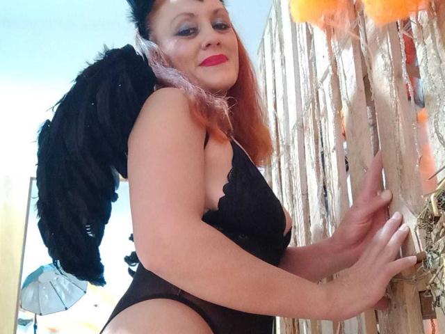 BRIGITH05 with black angel wings, pink hair, red lipstick, black lace lingerie, standing by a wooden fence, smiling, hands on the fence, showing her ass.