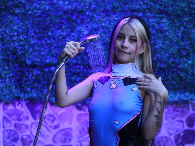 Yliria with blonde hair, blue and pink bodysuit, holding a showerhead, water on her tits, tattoo on right arm, smiling, blue and purple lighting, water droplets.