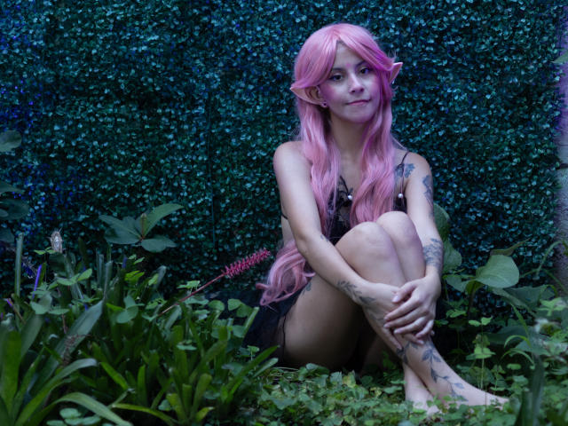 Yliria with pink hair, elf ears, tattoos, sitting cross-legged, naked, small tits, black shorts, forest floor, green plants, blue-green background, looking at camera, relaxed, cute.
