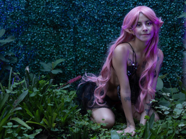 Yliria with pink hair, kneeling in greenery, wearing black lingerie, tattoos on arms, looking at camera, purple light on right side, ass slightly visible, long pink hair, cute.