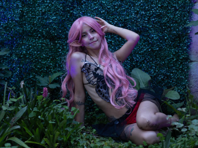Yliria with pink hair, elf ears, black lace top, red and black skirt, sitting in greenery, hand in hair, smiling, tattoos on arm and thigh, cute, sexy.