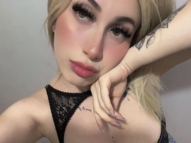 SammyViolet with blonde hair, pink blush, and pink lips, wearing black lace, has long eyelashes, nose ring, tattoos, and is resting her chin on her hand, showing her tits.