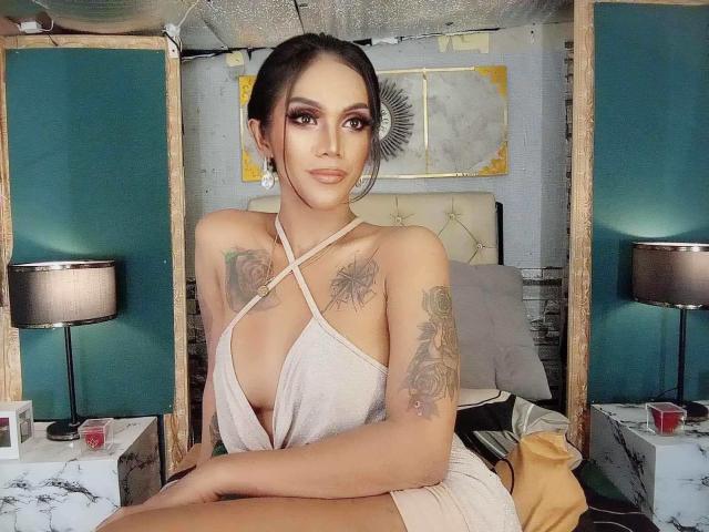 XTREMELYBeautyEmpire with tattoos on her arms and chest, wearing a revealing, criss-cross halter top, sitting on a bed, looking directly at the camera, with a neutral expression.