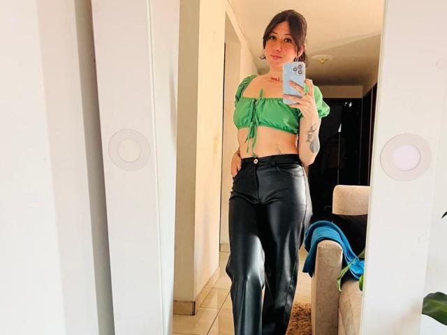 Valeriaaf with green crop top showing tits, black leather pants, tattoos, and a smartphone mirror selfie, standing in a hallway.