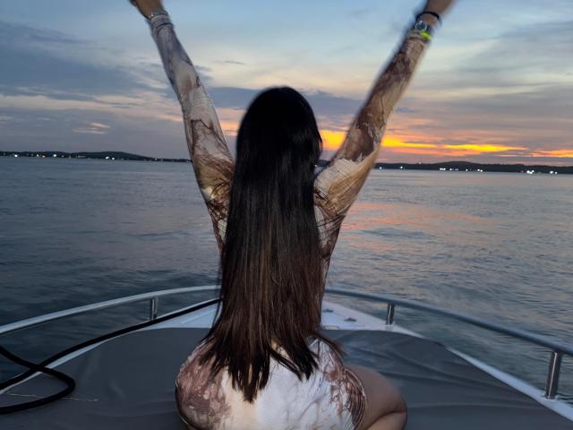 Heyly est assis sur un bateau, bras levés, dos face à la caméra, coucher de soleil sur l'eau, cheveux bruns mouillés.