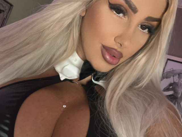 Close-up of TanyaMonroe. She has long, straight platinum blonde hair, thick, arched eyebrows, and heavily applied makeup with dark eyeshadow and glossy pink lipstick. She's wearing a black, cleavage-revealing outfit with a white collar, a small silver necklace is visible. Her skin is tan, and her eyes are slightly upward. The image focuses on her upper body, with her chest prominently in the frame. Her expression is confident, and she's looking at the camera. The overall style is bold and glamorous.