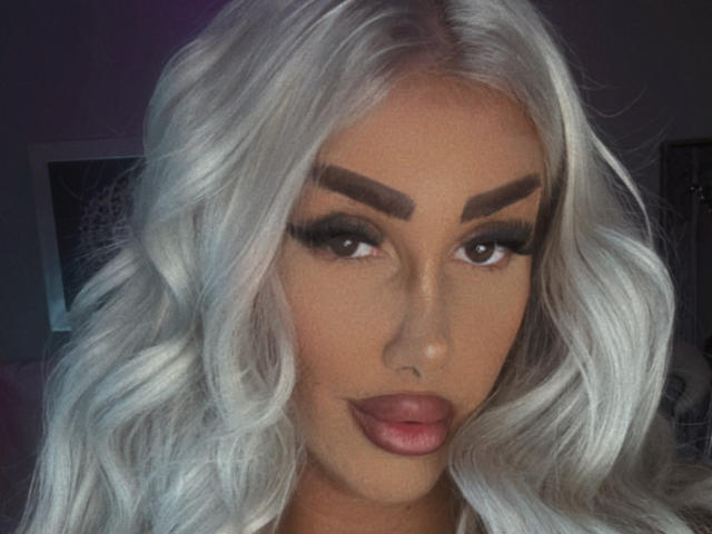 TanyaMonroe has platinum blonde, wavy hair, thick dark eyebrows, long eyelashes, and pink lips. Her tan skin is smooth, and she's staring directly at the camera.