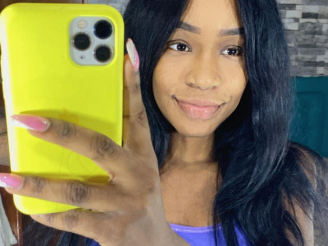 DannaSquirty holding a yellow phone, taking a selfie with pink nails, smiling at the camera, long black hair, brown skin, and cute face.
