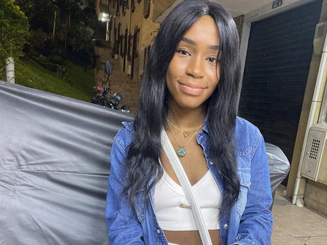 DannaSquirty with long black hair, dark skin, wearing a blue denim jacket over a white crop top, smiling softly, necklace with a green pendant, standing by a covered motorcycle.