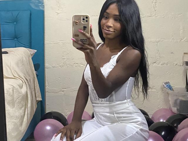 DannaSquirty with dark skin, long black hair, and small tits, wearing a white lace dress, taking a mirror selfie, surrounded by pink and black balloons.