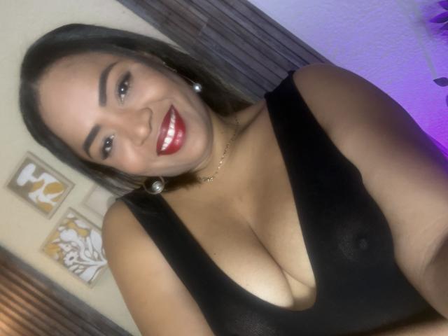 AliceBooty, avec ses cheveux foncés, son rouge à lèvres rouge et son haut noir transparent, dévoilant ses grandes seins, souriante, portant des boucles d'oreilles de perles et un collier en or. Arrière-plan intérieur flou..